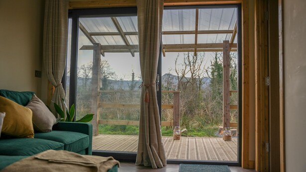 A room with a green sofa, a wooden deck outside, and a large glass door. - Cuilcagh Cabins (Enniskillen)