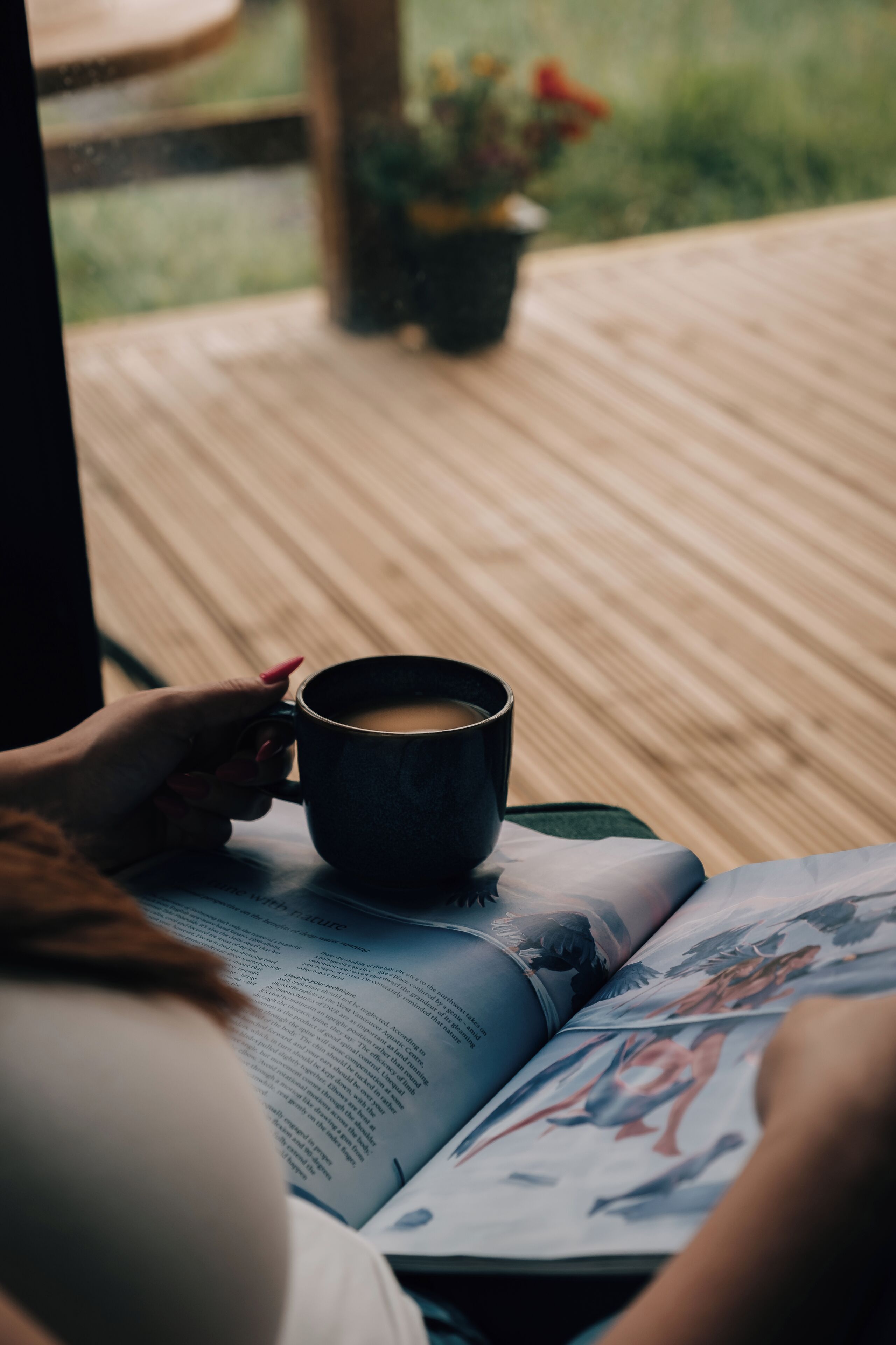 Uma pessoa lendo um livro com uma xícara de café em um deck de madeira.