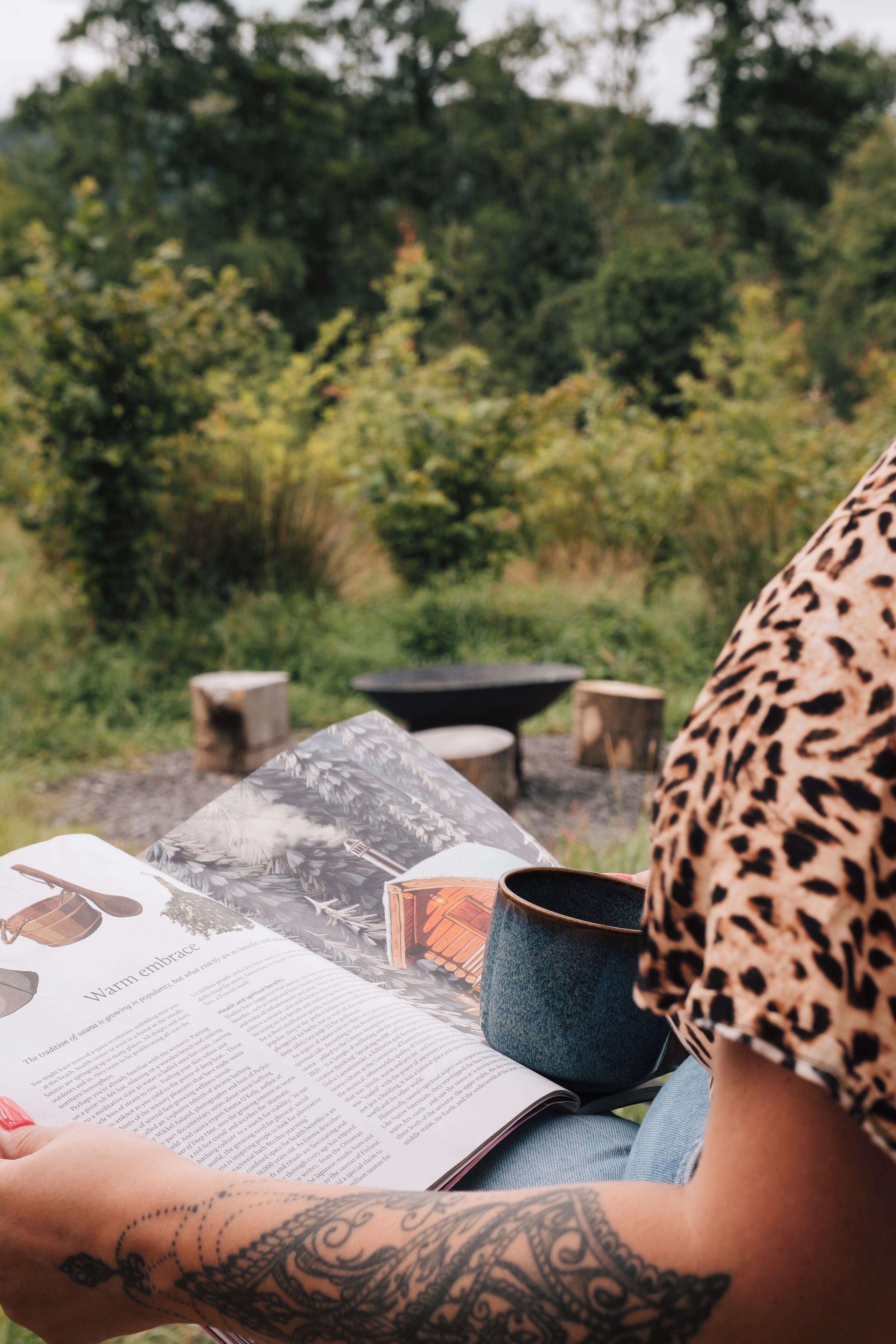 Una persona leyendo un libro al aire libre, con una mesa, una taza y un recipiente azul.