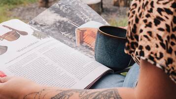 Una persona leyendo un libro al aire libre, con una mesa, una taza y un recipiente azul.