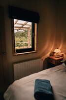 A bedroom with a window, a bedside table with a lamp, a radiator, and a bed with a blue towel.