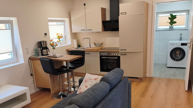 A modern kitchen with a built-in oven, a dining area with a table and chairs, and a grey sofa in a living area. - Home Sweet Home - Feel-good vacation home (Cuxhaven)