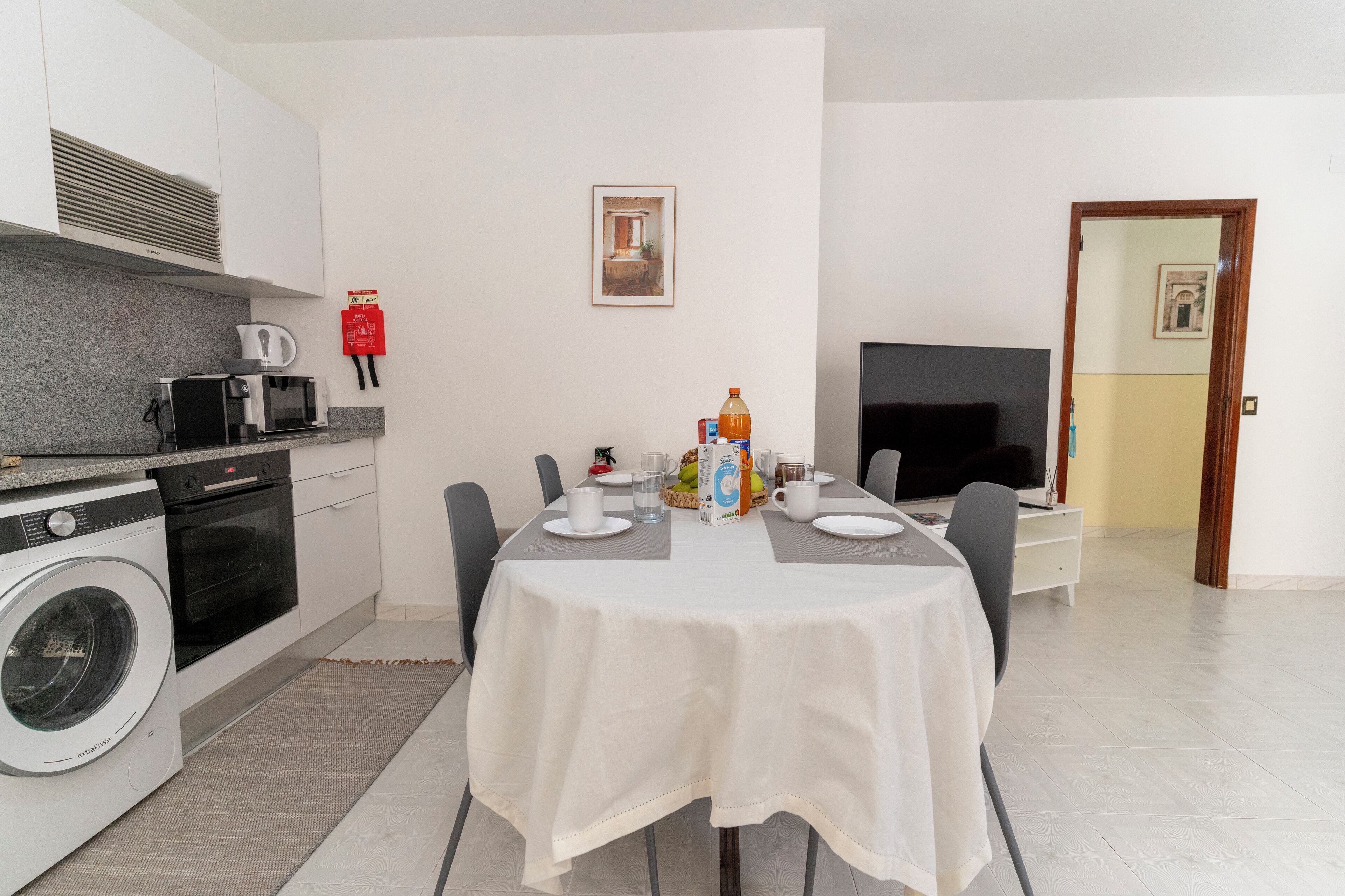 A modern kitchen with a microwave, toaster, and oven, a dining table set for four, and a TV in the living area.