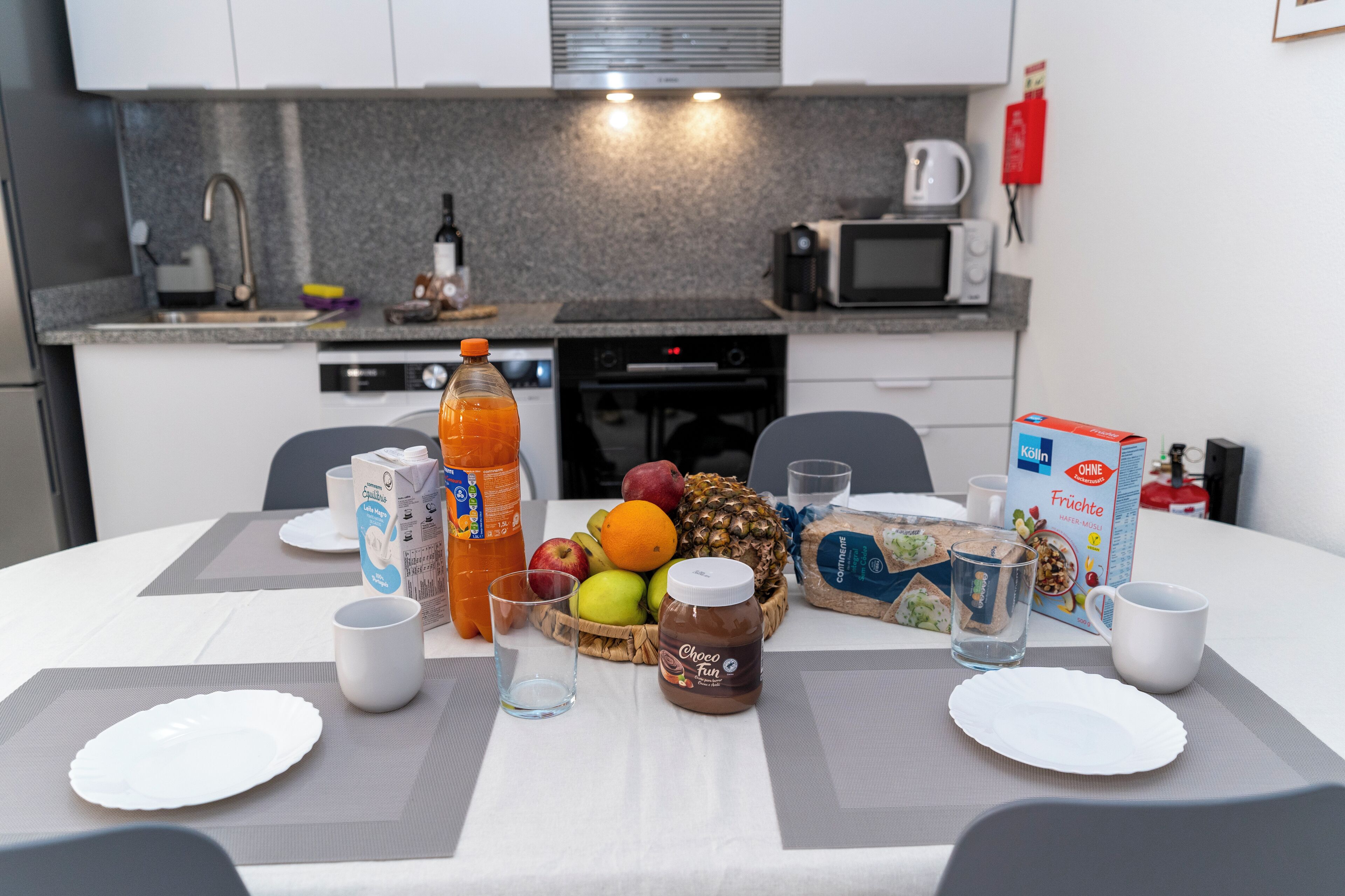 En morgenmadsopstilling med en skål frugt, juice og cornflakes på et bord i et moderne køkken.