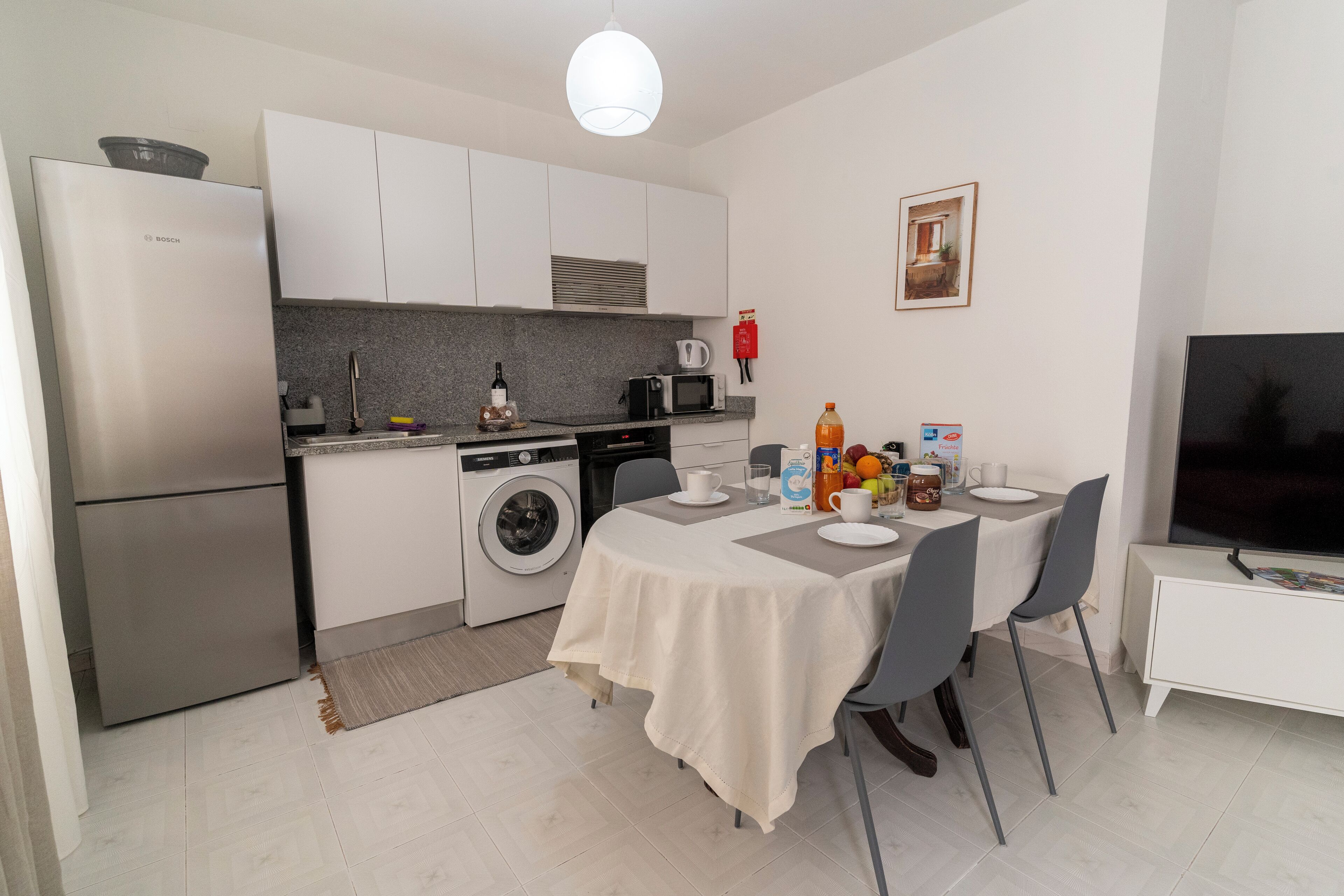 A compact kitchen with a Bosch refrigerator, a washing machine, a microwave, and a coffee maker. There is a dining table with chairs and a TV on a stand.