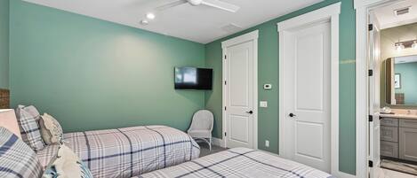 Quarto com duas camas, um ventilador de teto, uma TV e uma porta que dá para um banheiro.