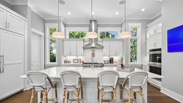 Una cocina moderna con armarios blancos, una isla grande con taburetes altos, iluminación colgante y vistas a espacios verdes al exterior.