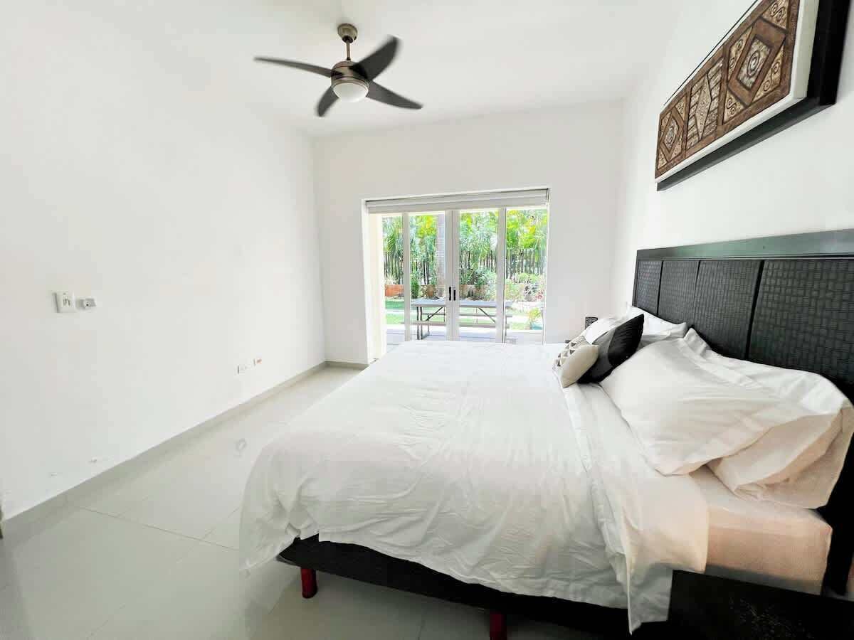 A bedroom with a bed, a ceiling fan, a large window, and a view of greenery outside.