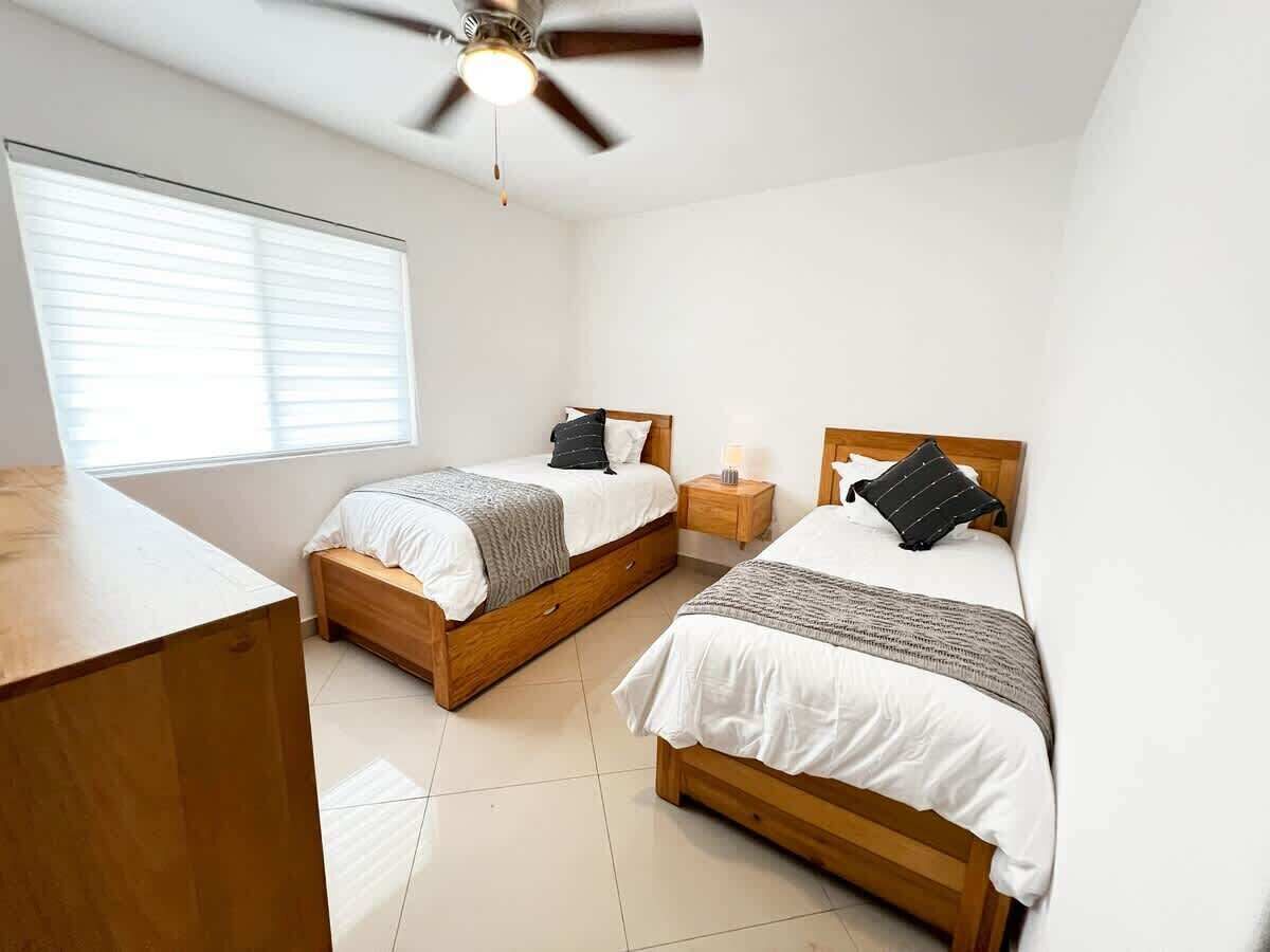Un dormitorio con dos camas de madera, un ventilador de techo, una mesita de noche con lámpara y una ventana con persianas.
