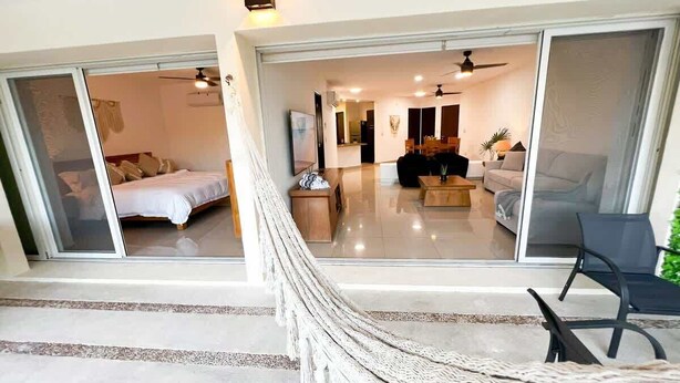A modern interior with a hammock, a bed, and a dining area visible through glass doors. - Beachside Condo w/ Private Terrace & 2 Pools (Puerto Aventuras)