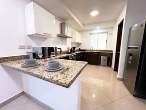 A modern kitchen with white cabinets, granite countertops, stainless steel appliances, and a small island with diningware. - Beachside Condo w/ Private Terrace & 2 Pools (Puerto Aventuras)