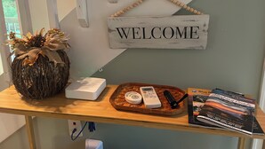 Una mesa de madera con un cartel que dice 'WELCOME', una planta decorativa, un dispositivo blanco, una bandeja tejida con un control remoto y una cámara, y una guía para visitantes.
