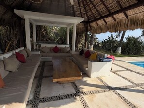 Ein überdachter Außenbereich mit einer Sitzecke, inklusive Sofa und Couchtisch, umgeben von einem Pool und tropischen Pflanzen.