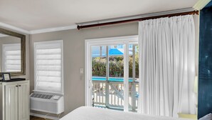 Un dormitorio con una cama grande, una ventana con cortinas blancas, vista a una piscina y un balcón con barandilla.