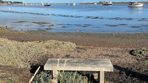 A wooden bench on a pebbly shore with boats on the water in the background. - Orford Holiday Cottage (Orford)