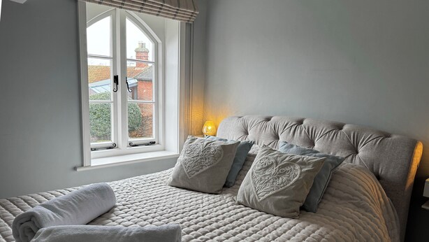 A bedroom with a large bed, a window with curtains, and a bedside lamp. - Orford Holiday Cottage (Orford)
