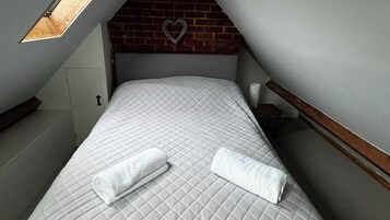 Un dormitorio con techo inclinado, una colcha acolchada, dos toallas enrolladas, una decoración con forma de corazón en la pared y una mesita de noche con una lámpara.
