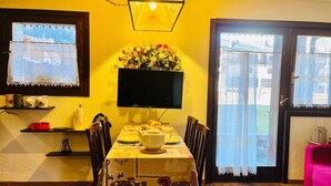 Un área de comedor con una mesa dispuesta para dos personas, un televisor montado encima y una ventana con cortinas de encaje.