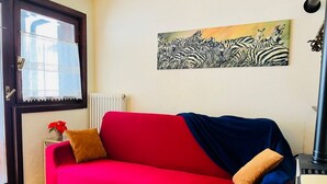 Un sofá rojo con dos almohadones, un radiador, una ventana con cortinas de encaje y un cuadro de una cebra en la pared.