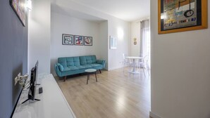 Een woonkamer met een blauwgroene bank, een kleine witte tafel en een eethoek met transparante stoelen. Er hangen kunstwerken in lijsten aan de muren en een poster aan de rechterkant.