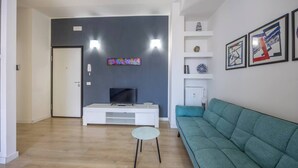 Een moderne woonkamer met een flatscreen televisie op een witte televisiemeubel, een blauwgroene bank en een kleine ronde tafel.