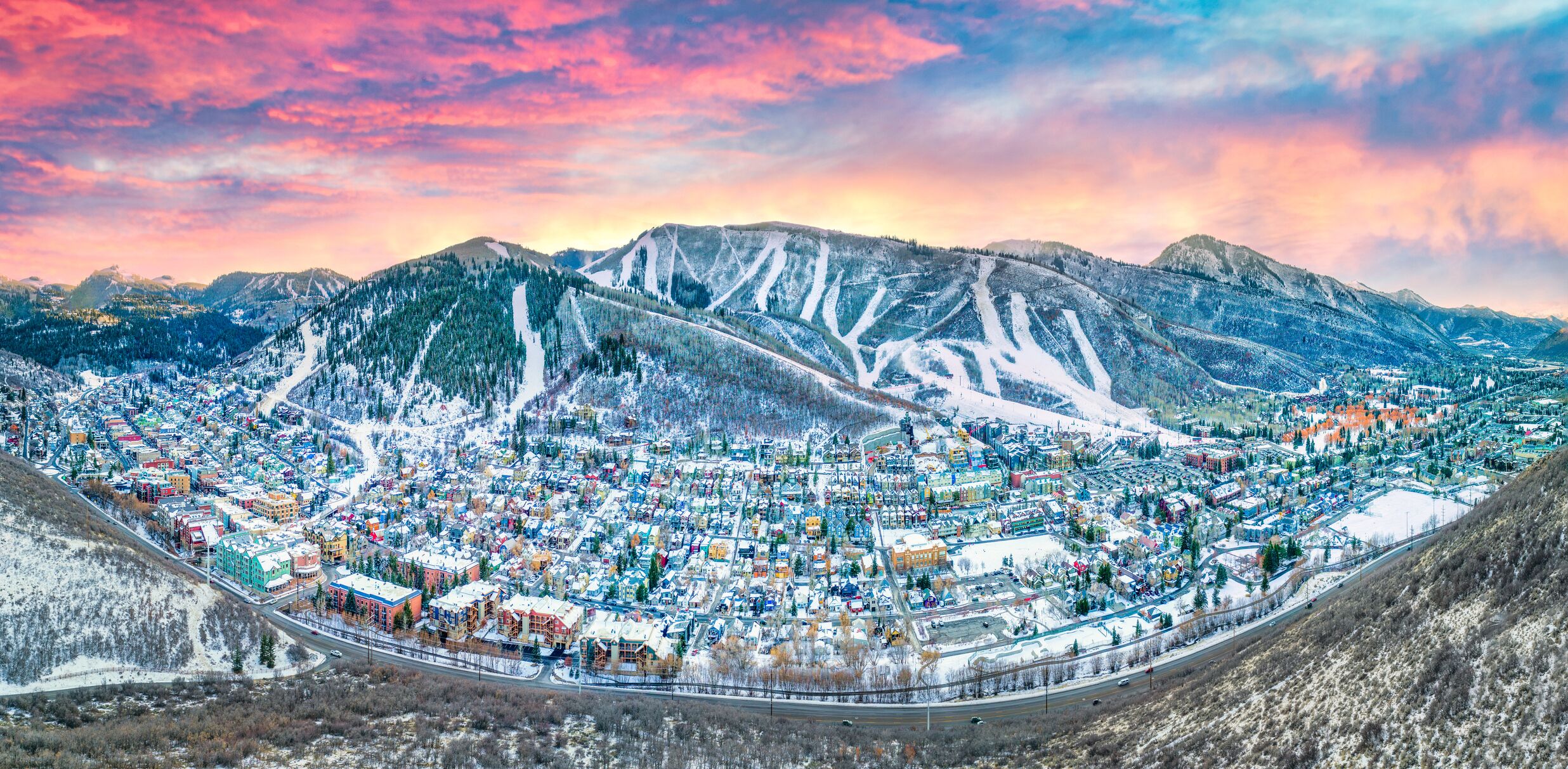 A panoramic view of a town with a mountain backdrop, featuring a mix of residential and commercial buildings, roads, and a railway track.