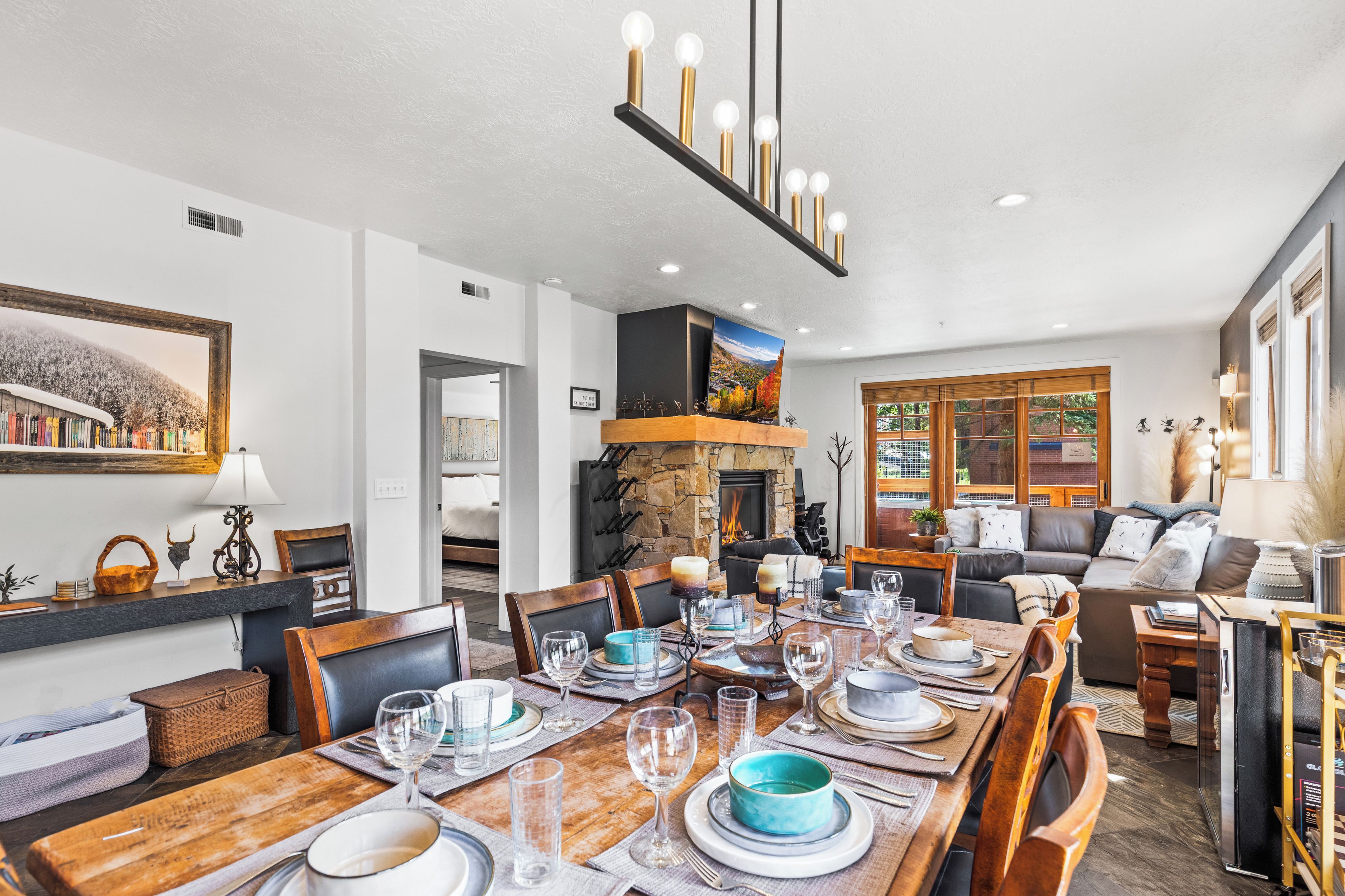 Un comedor con mesa de madera dispuesta para una comida, una chimenea de piedra y una sala de estar con sofá y televisor.