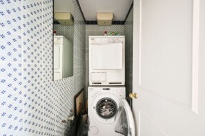 A compact laundry area with a washing machine and dryer, a small cabinet, and patterned wall tiles. - Argyl | Stanhope Gardens: Bright 2 bedroom Apt. (London)