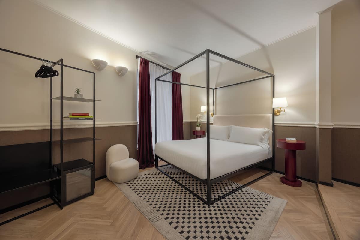 A modern hotel room with a four-poster bed, a small seating area, a bookshelf, and a bedside table with a lamp.