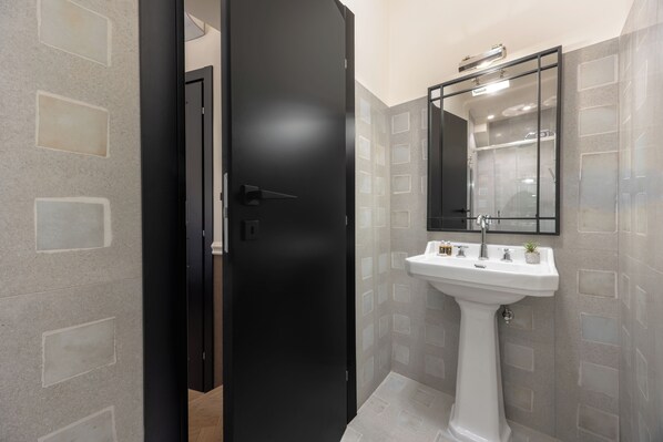 Une salle de bain avec un lavabo sur colonne blanc, un miroir au-dessus, et une porte donnant sur une autre pièce.