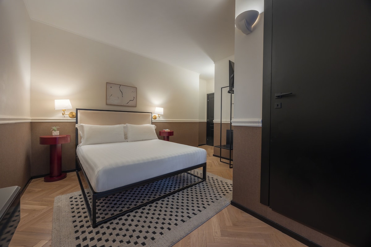 A modern hotel room with a large bed, two bedside tables with lamps, a wall-mounted TV, and a wardrobe.