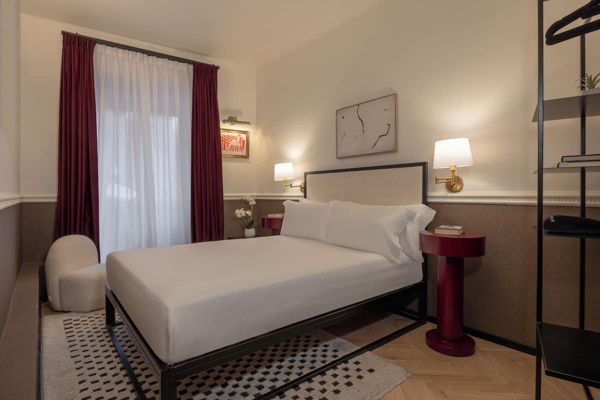 A hotel room with a bed, a chair, two bedside tables with lamps, a wall clock, and a shelf with a plant.