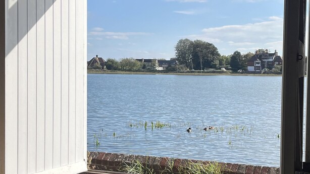 Vista de un lago con casas al fondo, cielo despejado y una estructura de madera en primer plano.