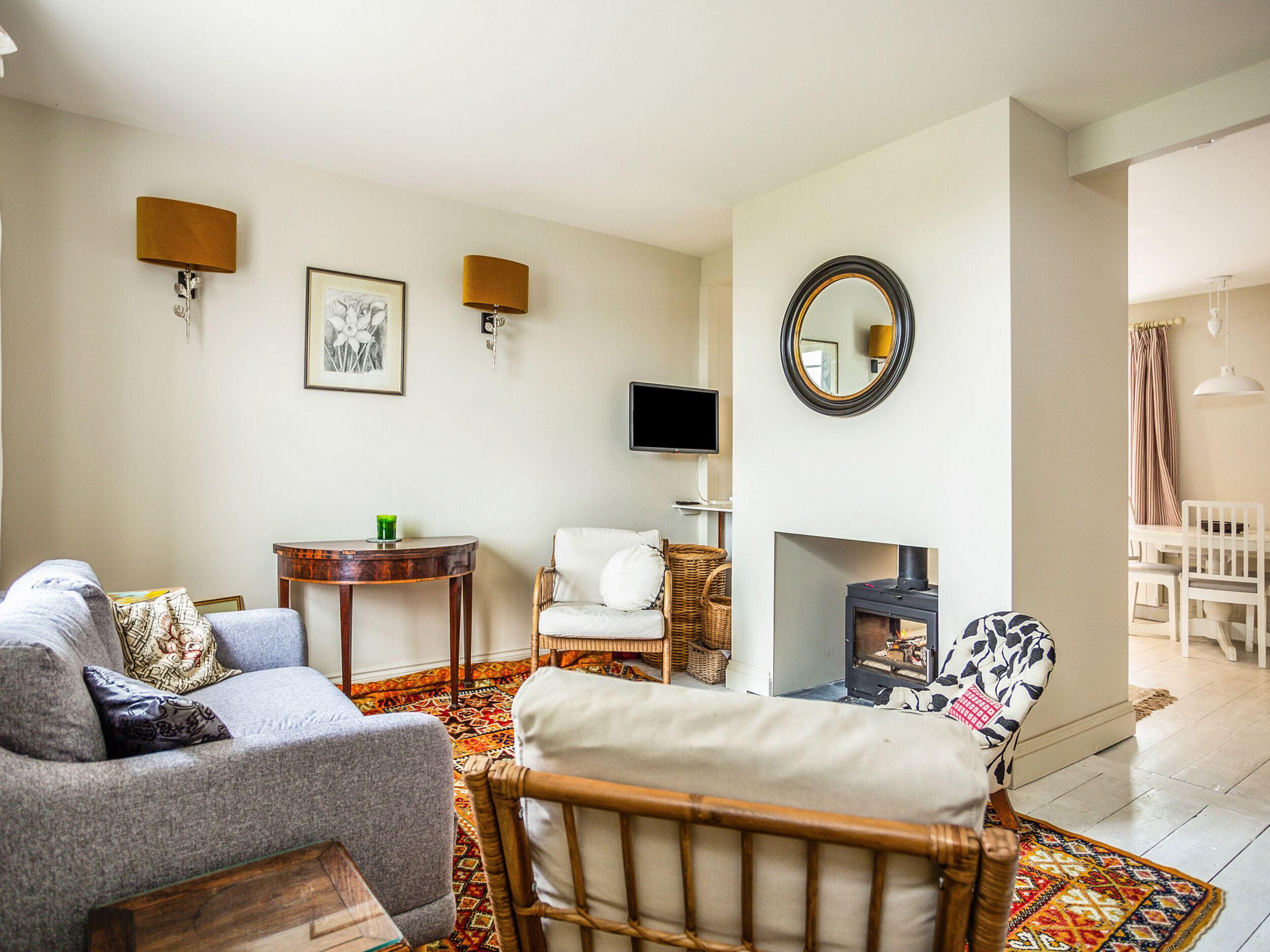 A living room with a sofa, armchair, and a small table with a lamp. There is a fireplace with a TV above it and a mirror on the wall.