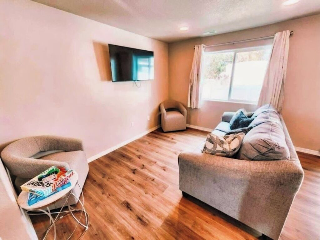 Un salon comprenant un canapé, un fauteuil, une petite table avec des magazines, un téléviseur à écran plat fixé au mur et une fenêtre avec des rideaux.