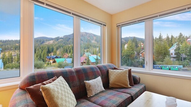 A room with a sofa facing large windows offering a view of a mountainous landscape and a lake. - The Haven at Blueberry Hill - Mount Washington ski-in/out (Comox-Strathcona C)