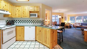 A modern kitchen with wooden cabinets, a microwave, and a dishwasher. There is a dining area with a table and chairs, and a living room with a sofa and a window. - The Haven at Blueberry Hill - Mount Washington ski-in/out (Comox-Strathcona C)