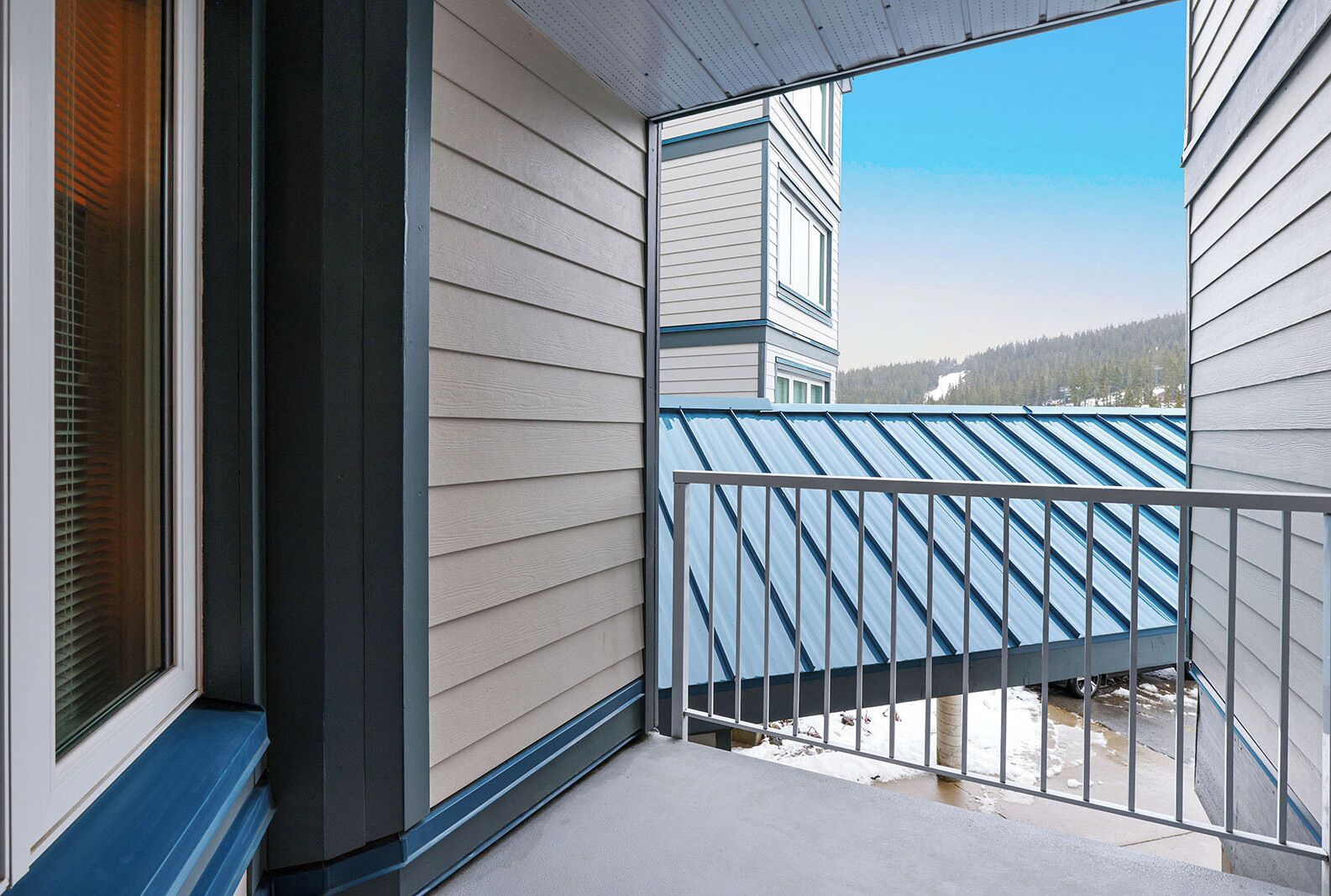 Een balkon met een metalen hekwerk, beige gevelbekleding en uitzicht op een gebouw met een blauw dak.