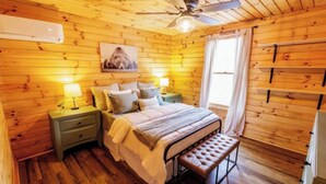 A wooden cabin-style bedroom with a bed, nightstands, a bench, a ceiling fan, and a window with curtains. - Secluded 2 Bedroom Timber Ridge Cabin- Hot Tub, Lake Boat Ramp 1 Mile, Fishing (Louisa)