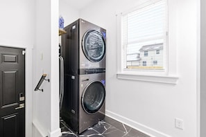 Une buanderie équipée d’une machine à laver et d’un sèche-linge superposés, d’une porte et d’une fenêtre avec des stores.