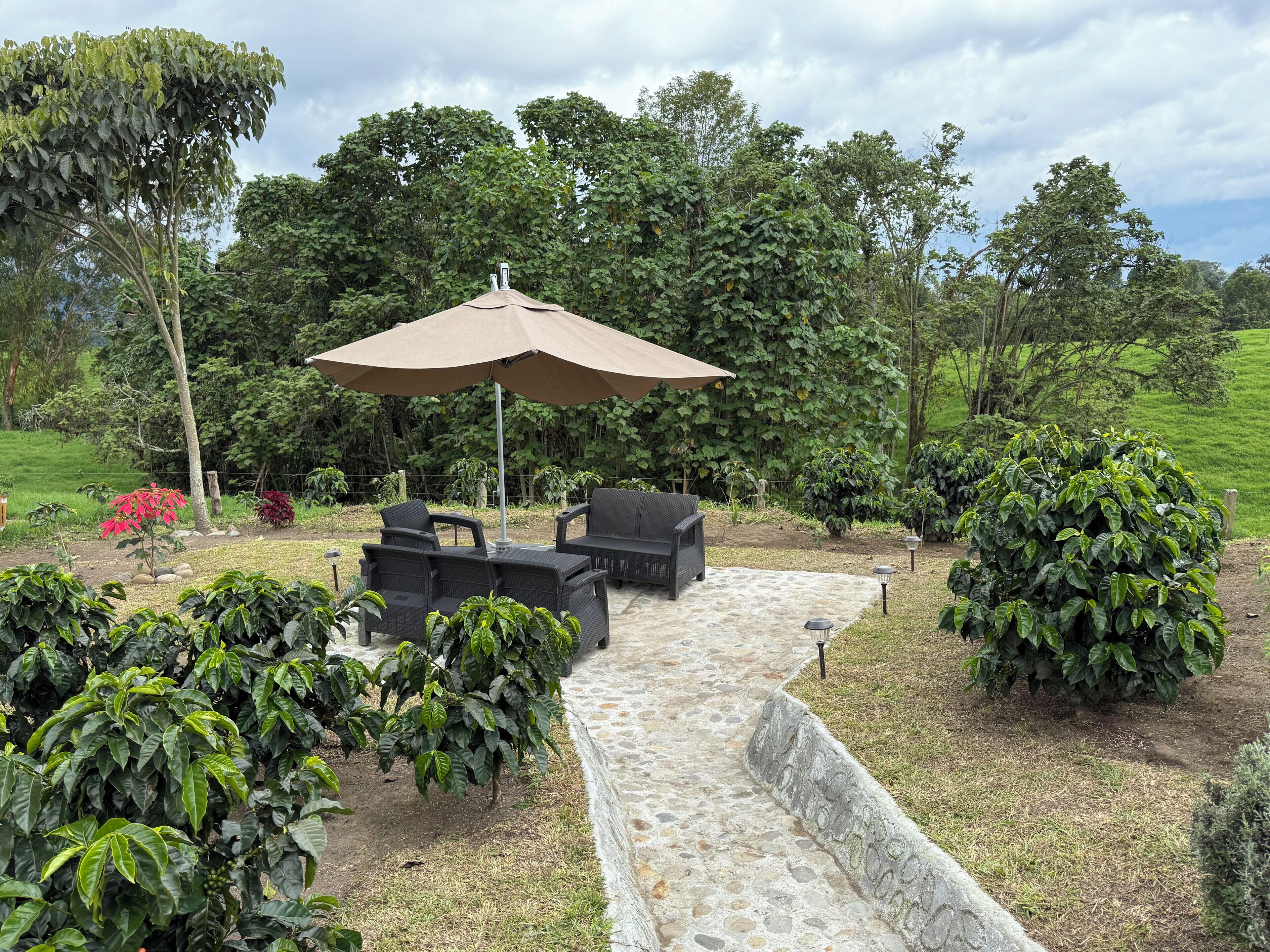 An outdoor patio area with a stone pathway, two black wicker chairs, a large beige umbrella, and lush greenery in the background.