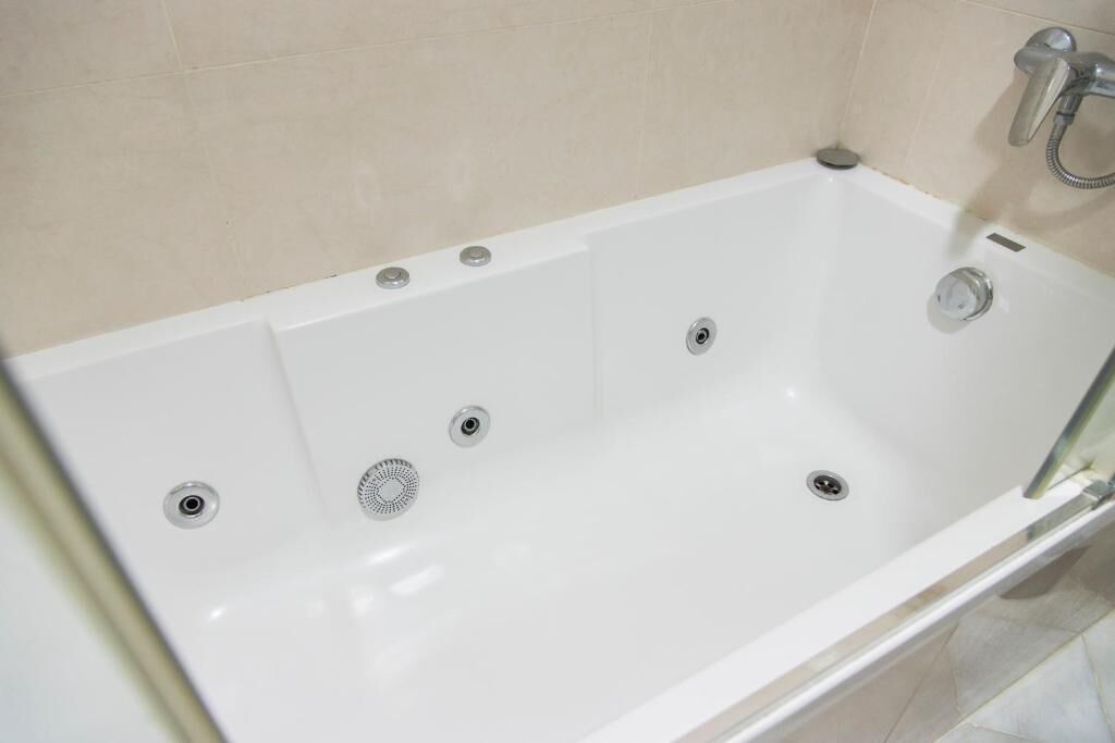Eine weiße Badewanne mit mehreren Abläufen, einer Handbrause und einer gläsernen Duschkabine.