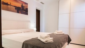 A hotel room with a large bed, a painting of a sunset, a wardrobe, and a neatly made bed with towels. - Night In Sevilla (Seville)