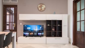 A living room with a flat-screen TV on a white entertainment unit, a clock on the wall, and a door with a glass panel. - Night In Sevilla (Seville)