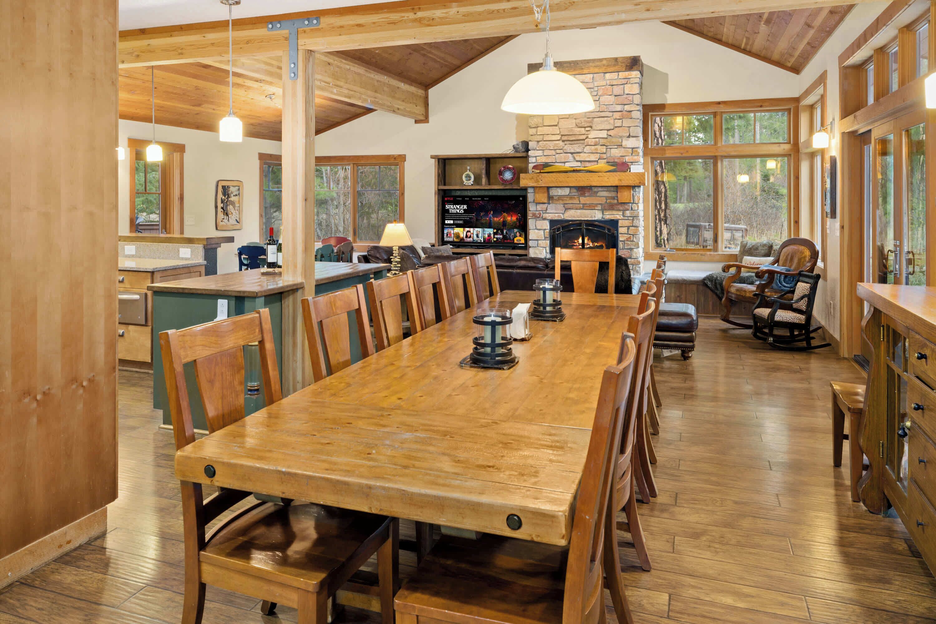 A spacious dining area with a long wooden table and chairs, a stone fireplace, and large windows offering views of the outdoors.