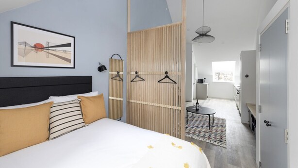 Una habitación de hotel moderna con una cama, un separador de madera, una zona de comedor y vistas al exterior.