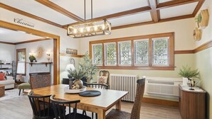 Ein Essbereich mit Holztisch, vier Stühlen und einer Hängeleuchte. Ein Fenster mit Jalousien, ein Heizkörper und eine Pflanze auf einem Beistelltisch sind ebenfalls vorhanden.