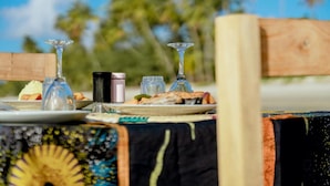 Une table dressée avec des assiettes remplies de nourriture, des verres et un thermos, sur une plage où l’on aperçoit en arrière-plan des chaises en bois et des palmiers.