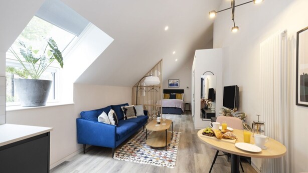 Um apartamento moderno com teto estilo loft, um sofá azul, uma mesa de jantar com café da manhã e uma grande janela com uma planta.