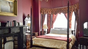 Ein Bett mit Baldachin, eine rote Bettdecke, ein Nachttisch mit Lampe, eine Kommode mit Büchern und ein Gemälde an der Wand.